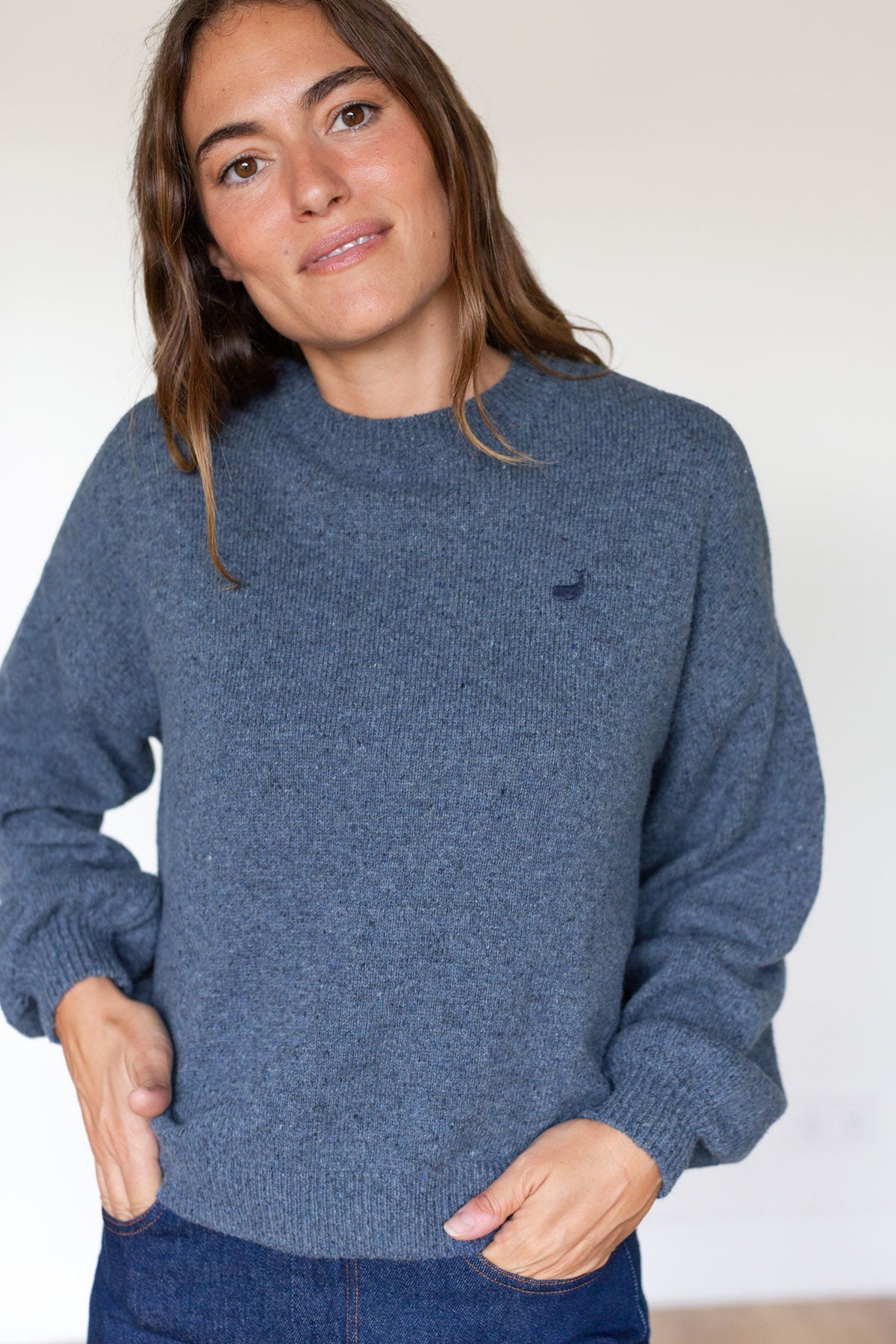 Woman with hands in pockets wearing a textured blue knit sweater with a small embroidered whale.