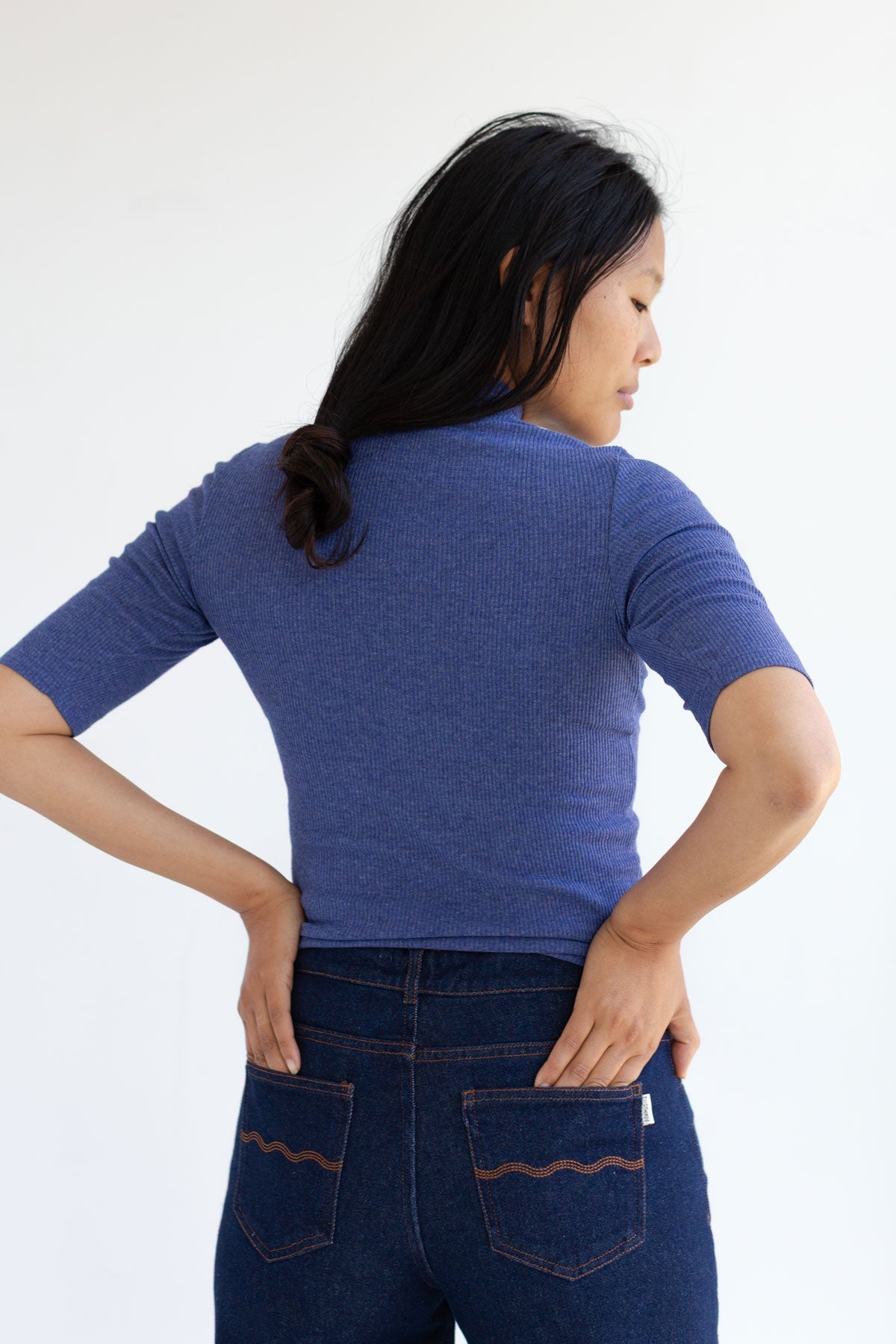Back view detail of a woman wearing a slim-fit blue top and dark blue jeans with her hands in the back pockets.