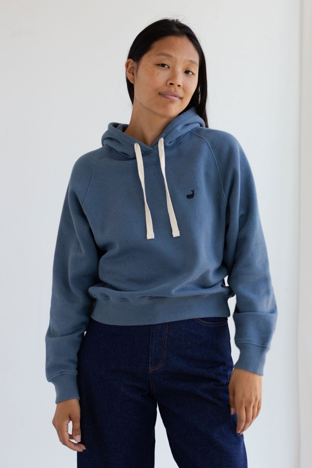Woman wearing a dusty blue raglan hoodie with a small embroidered whale.