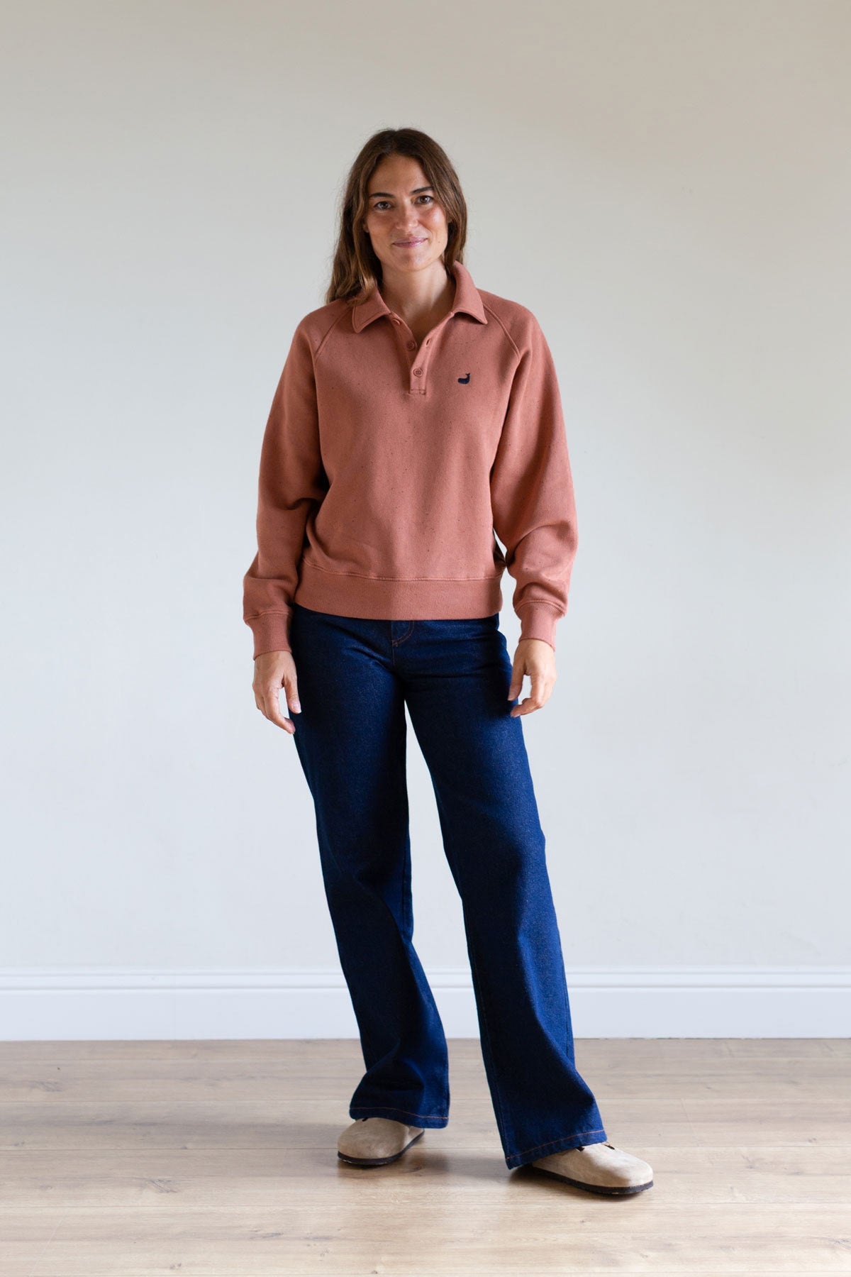 Full body front view of a woman wearing an adobe rose polo sweatshirt and blue jeans.