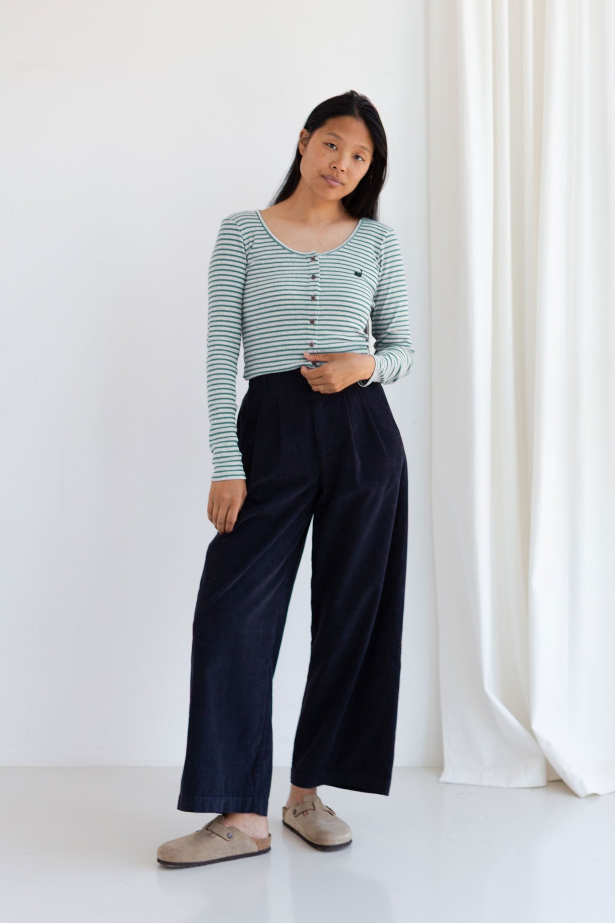 Full body front view of a woman wearing an alga green striped long sleeve top with buttons and wide-leg corduroy pants.