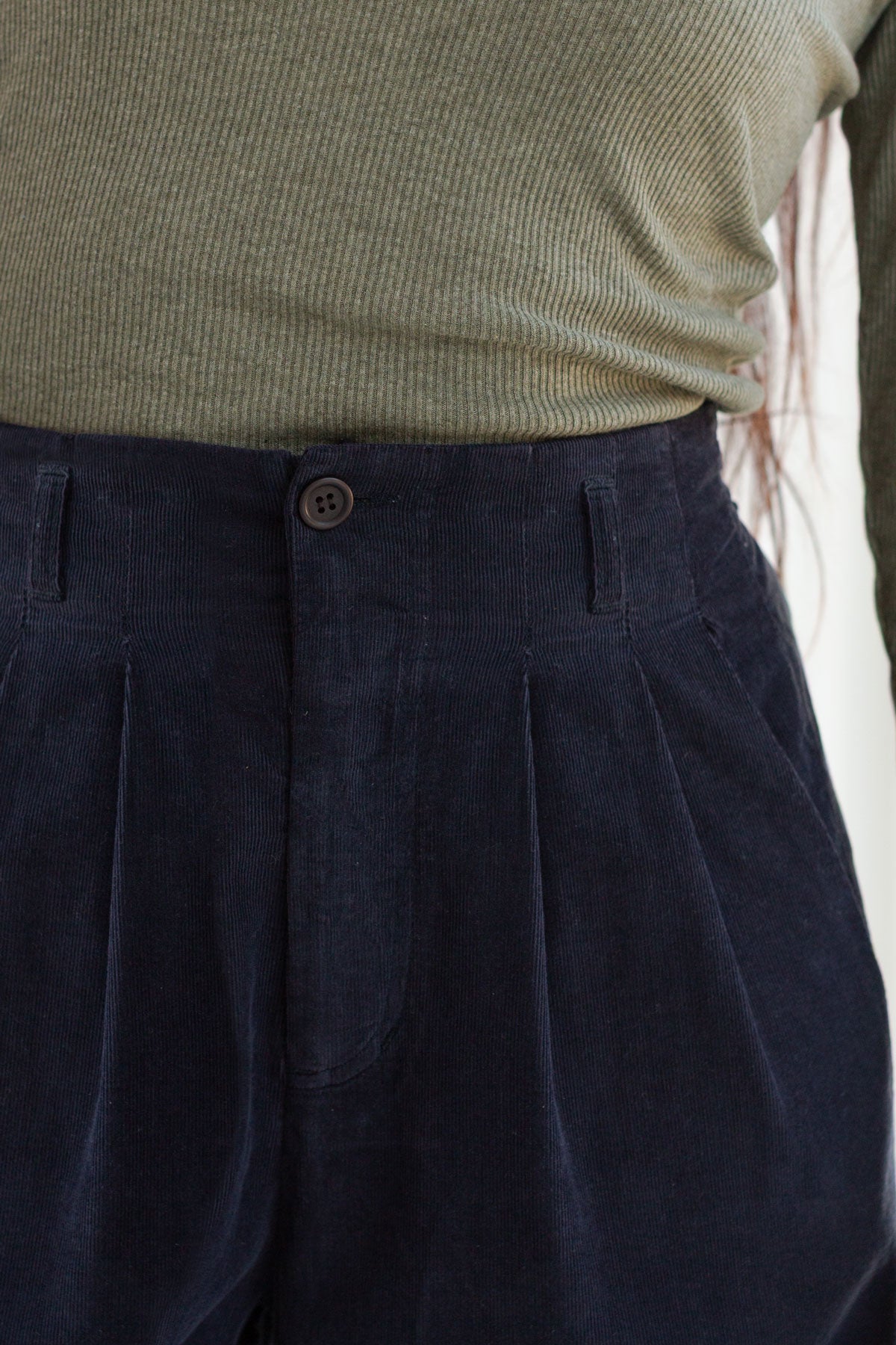 Close-up front view of the waist, pleats, and button closure on midnight blue micro corduroy pants.