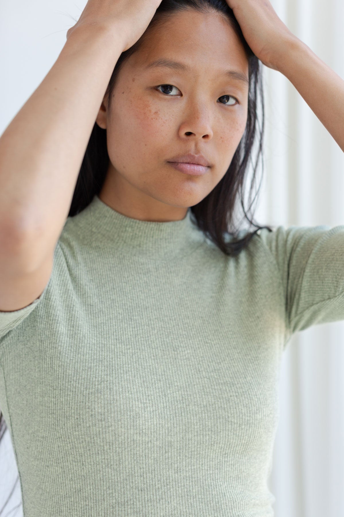 Close-up of a woman with hands on her head wearing a desert sage green mock-neck ribbed top.