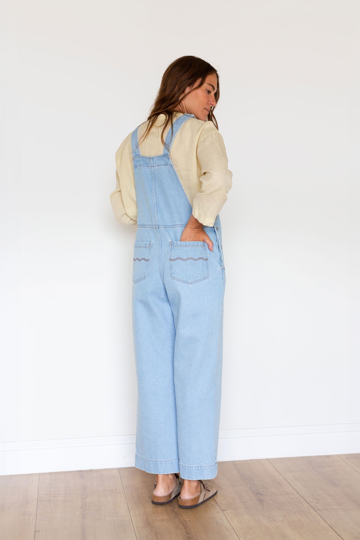 Back view of a woman wearing wide-leg light blue denim overalls with one hand in back pocket.