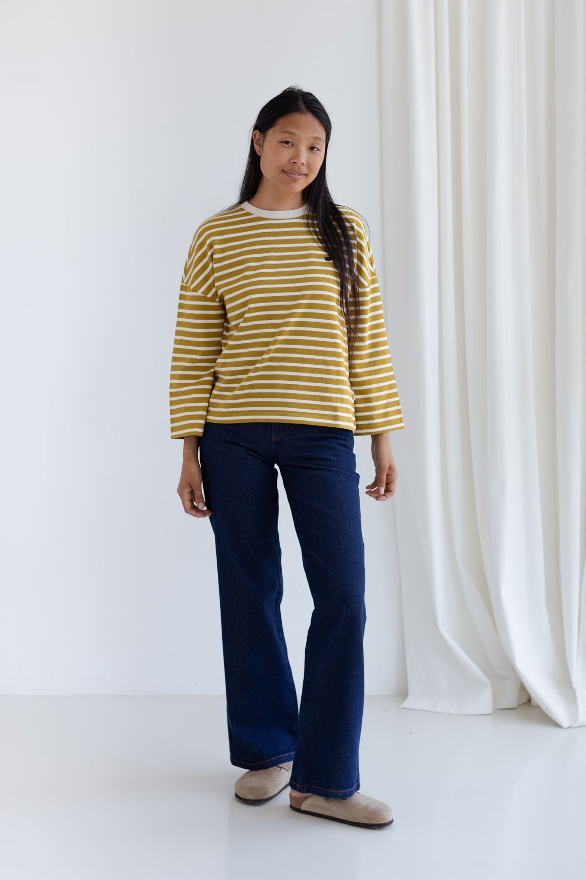 Full body view of a woman wearing a relaxed-fit mustard striped top and wide-leg dark blue jeans.