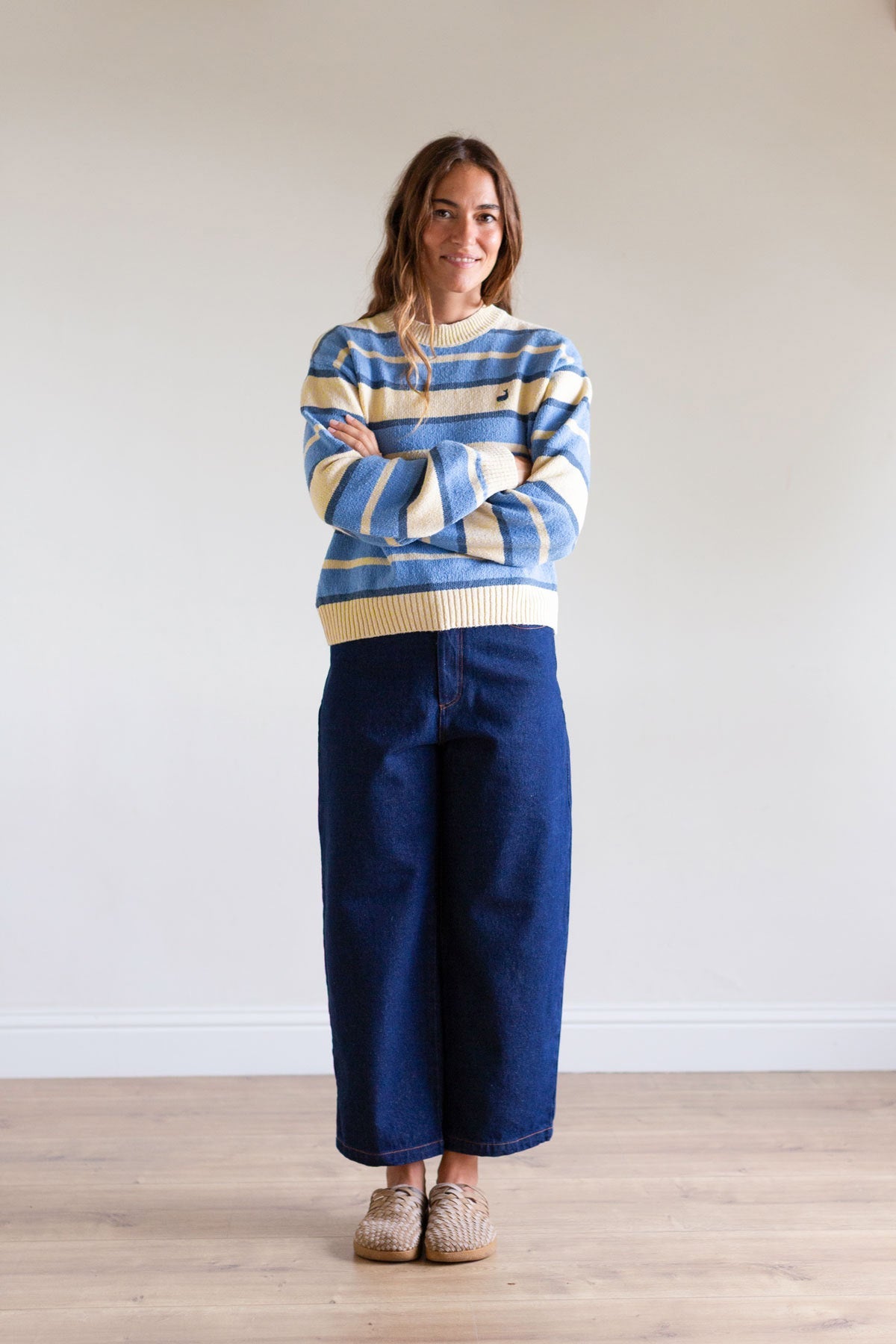 Full body shot of a woman with arms crossed wearing a blue striped sweater and wide jeans.