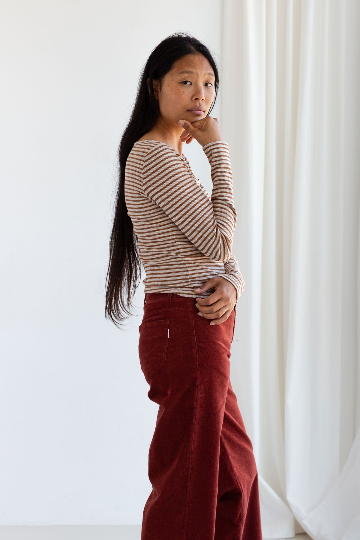Side profile view of a woman posing in a caramel striped ribbed top and red corduroy pants.