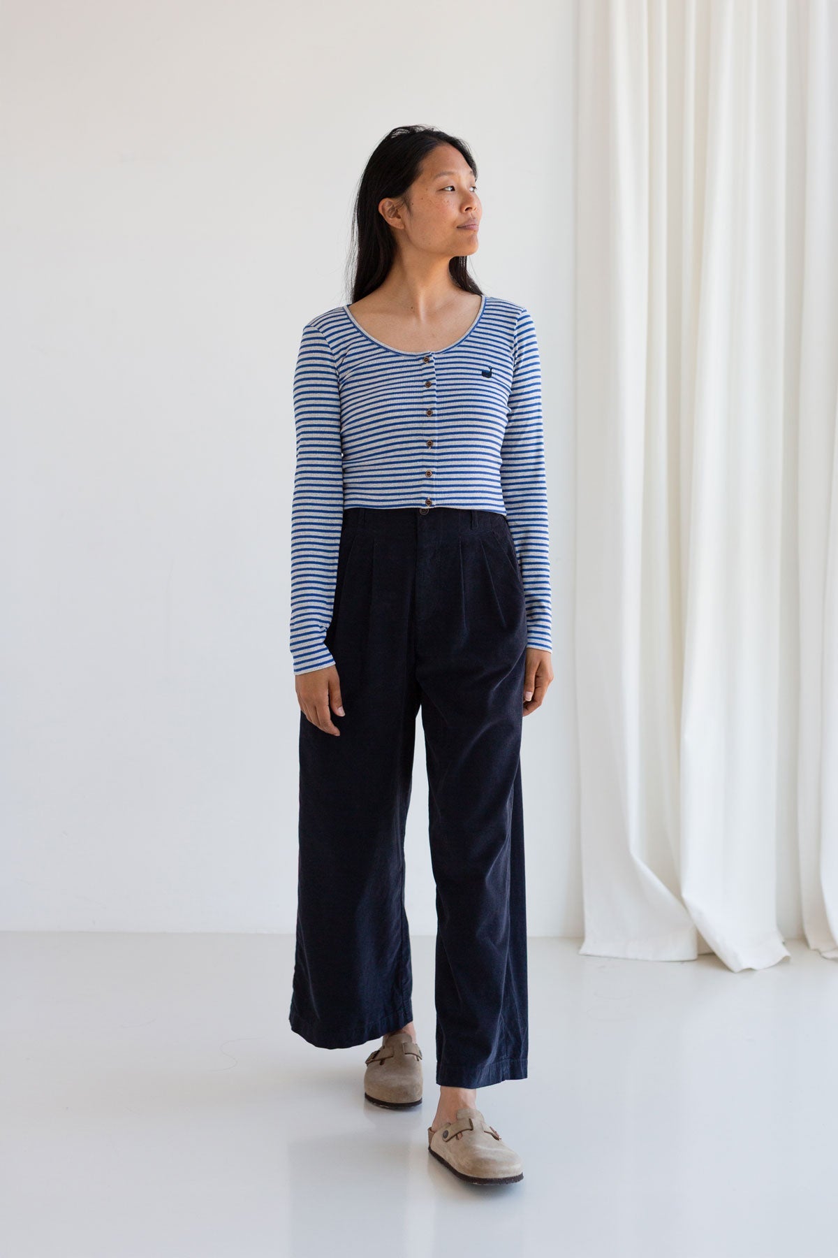 Full body front view of a woman wearing a blue striped top with buttons and dark wide-leg microcorduroy pants.