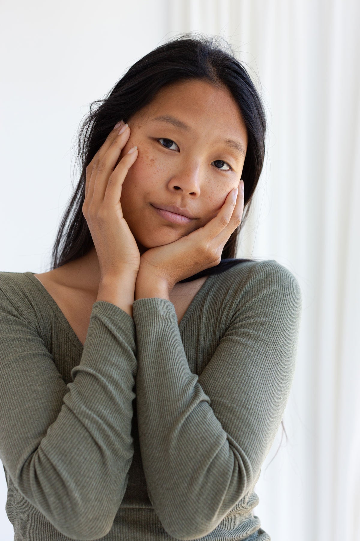 Woman with hands on her face wearing a ribbed olive green long sleeve top.