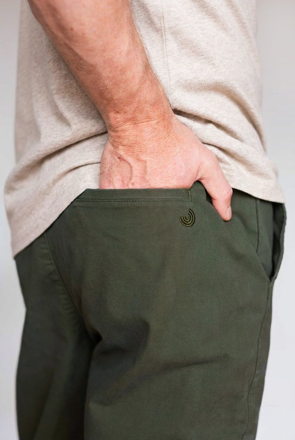 Hand in the back pocket of dark moss pants with a small embroidered logo detail.
