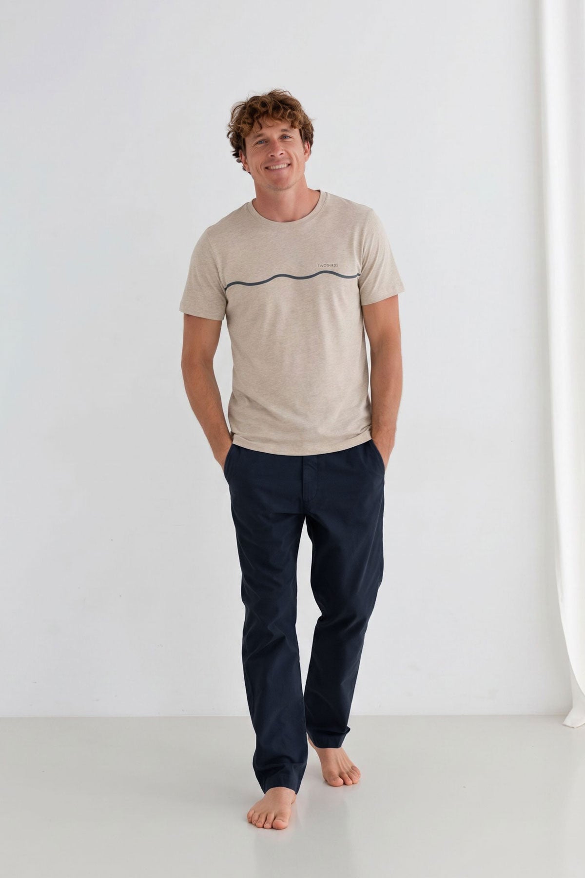 Man standing wearing a beige t-shirt and midnight blue pants with his hands in pockets.