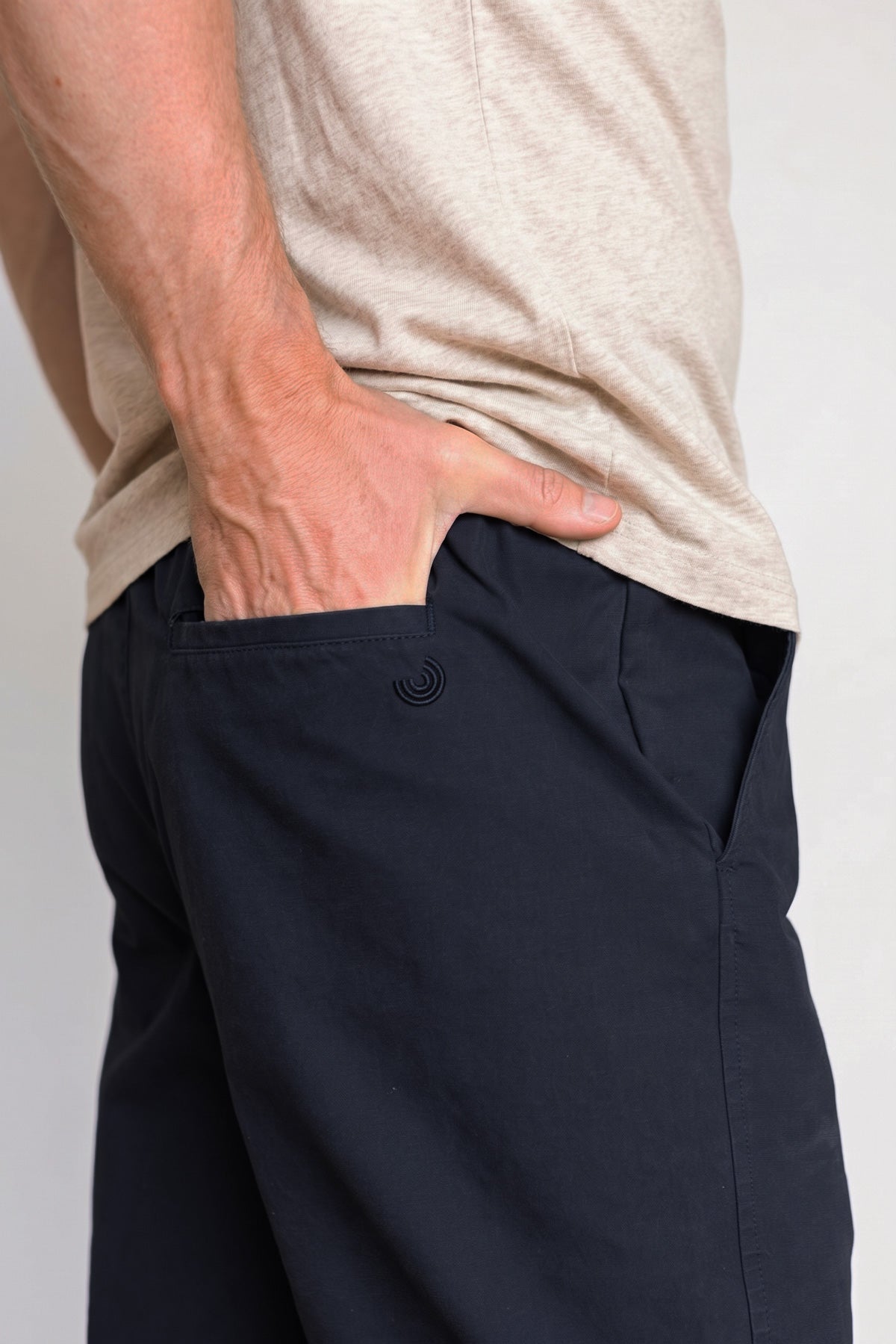 Close-up side view showing a hand in the back pocket of midnight blue pants with a small embroidered logo detail.